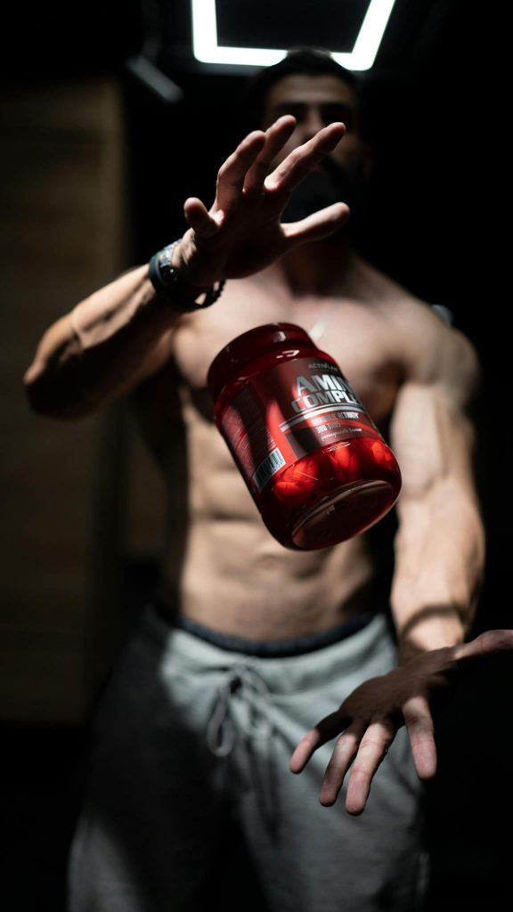 A shirtless man skillfully tosses a supplement jar in a dramatic indoor setting, showcasing fitness and agility.